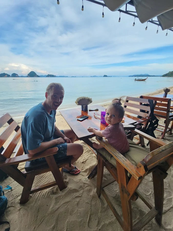 dad and baby sitting on the beach to have a meal in thailand smiling infront of a sunset