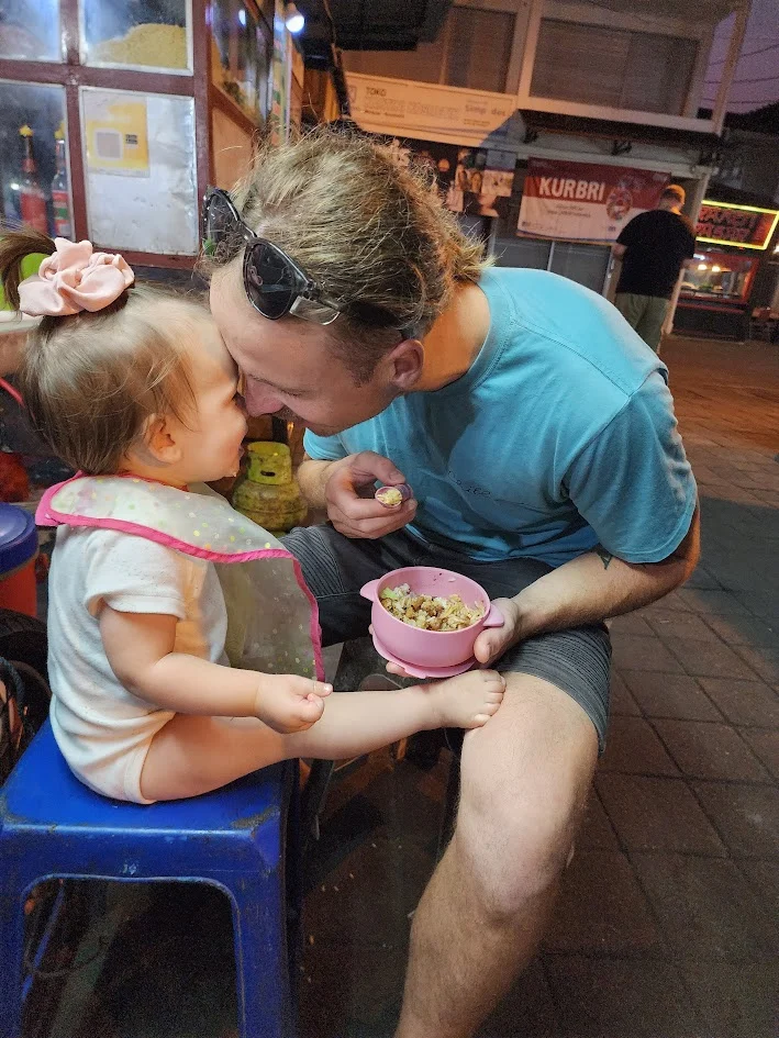 A dad sitting on a stool with his daughter at a food market in Bali. They’re eating dinner and smiling at each other.