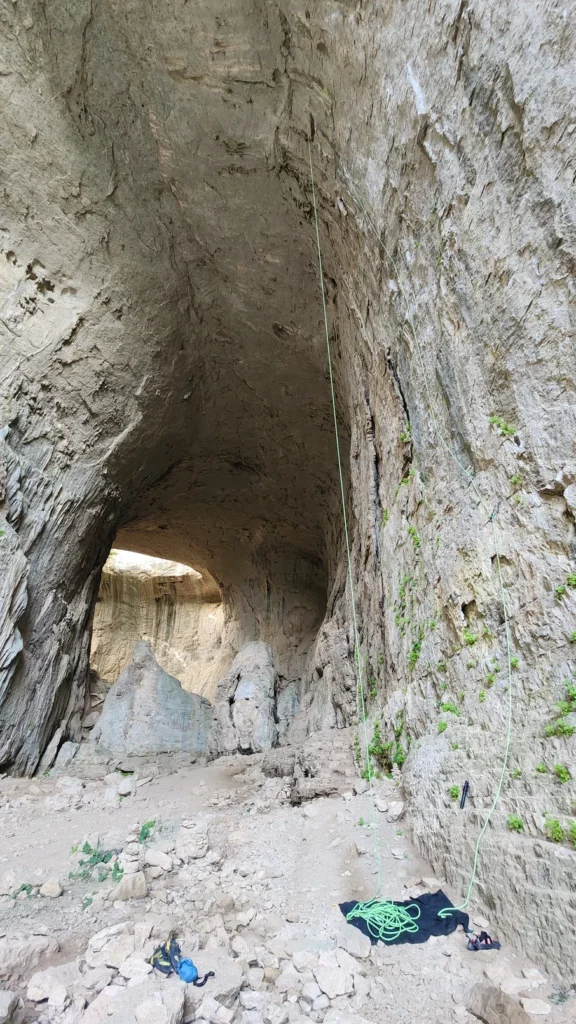 A climber on a steep route inside Prohodna Cave, with ropes and harness gear visible